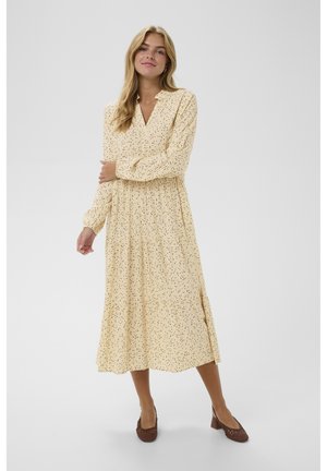 Lang, gul kjole med sort prikket mønster, plisseret design, V-udskæring og lange ærmer. Bæres med brune sko med lave hæle.