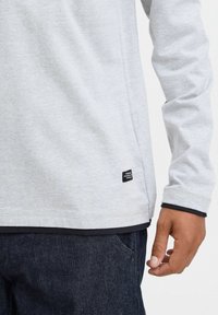 Hellgraues Langarmshirt aus weichem Stoff, mit einem schwarzen Etikett am Saum und einer blauen Schicht, die am Bund sichtbar ist.