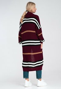Sweter kardigan w kolorze burgundowym z poziomymi paskami w kolorze czarnym, białym i beżowym. Posiada luźny krój i długie rękawy, noszony na niebieskich dżinsach.