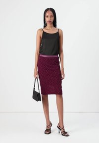Schwarzes Seidentop, strukturierter bordeauxroter Bleistiftrock mit floralem Muster und schwarze Riemchenheels. Model hält eine kleine schwarze Handtasche.