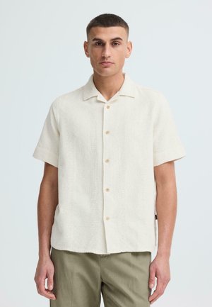 Jonge man met kort haar, gekleed in een crème kleuren overhemd met korte mouwen en olijfgroene broek, staand tegen een eenvoudige lichte achtergrond.