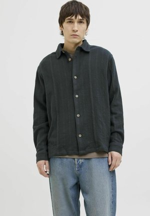 Chemise noire texturée avec un col, fermeture à boutons et manches longues. Associée à un jean en denim bleu clair. Fond neutre.
