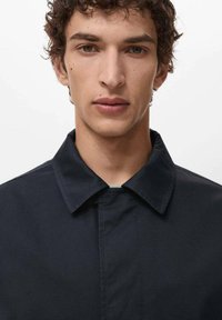 Giacca blu navy con colletto a punta e chiusura con bottoni. Il materiale appare liscio con una leggera lucentezza, caratterizzato da dettagli di design minimalisti.