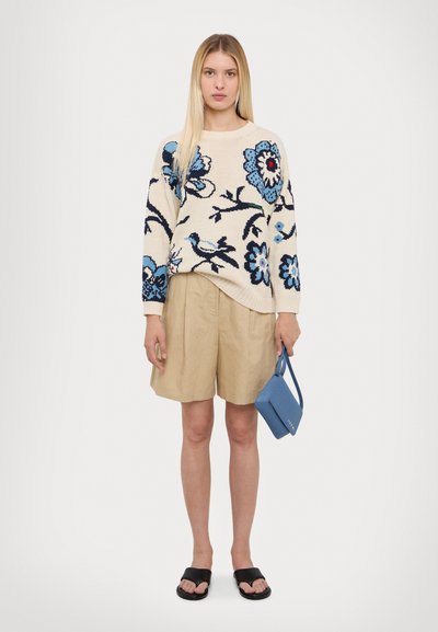 Suéter de punto con patrón floral en azul y negro sobre crema, combinado con pantalones cortos beige. Accesorizado con un pequeño bolso azul y sandalias negras.