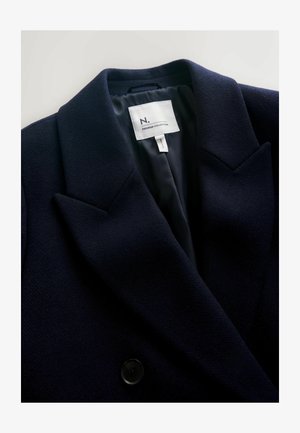 Tummansininen bleiseri, jossa on teksturoitu kangas, terävät kaulukset ja kaksinappinen kiinnitys. Sisäpuolella on sileä vuori ja brändilogo.