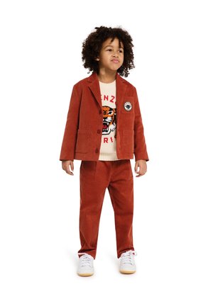Kind in een roestkleurig corduroy pak met een patch, gecombineerd met een sweater met een tijgerontwerp op een crèmekleurige achtergrond, witte sneakers.