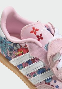 Zapatilla con estampado floral rosa que presenta una parte superior texturizada, diseño de tres franjas blancas y suela de goma; incluye la palabra "Samba" en dorado en el costado.