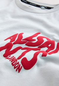 Hvid bomuld t-shirt med fed, struktureret rød tekst, der lyder "ALLSAINTS LONDON," med en klassisk rund halsudskæring.