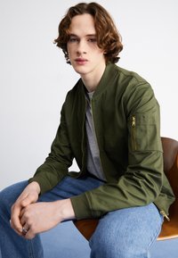 Veste bomber verte au tissu lisse, avec une poche zippée sur la manches, portée par-dessus un t-shirt gris et associée à un jean bleu.