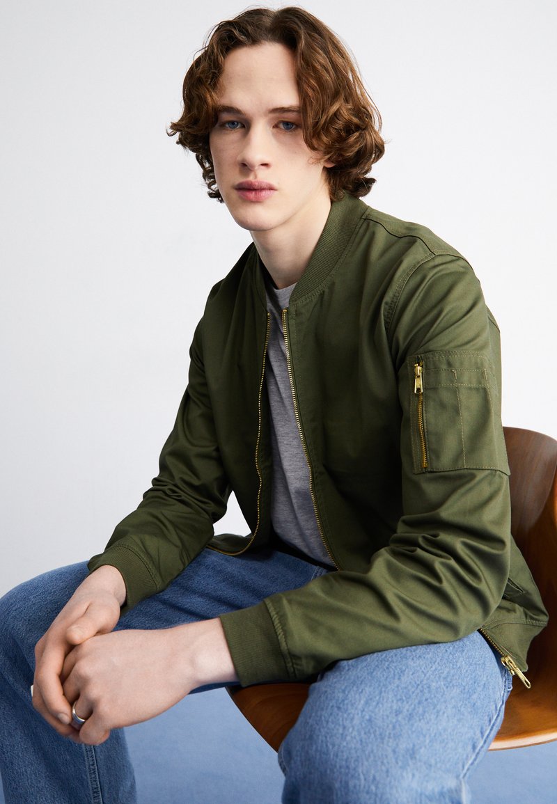 Veste bomber verte au tissu lisse, avec une poche zippée sur la manches, portée par-dessus un t-shirt gris et associée à un jean bleu.