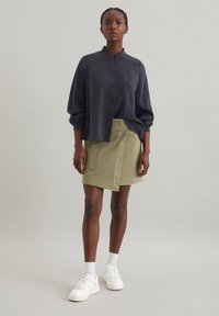 Camicia blu navy con colletto alto, abbinata a una gonna avvolgente verde oliva. Sneakers bianche completano l'outfit. Design semplice e raffinato.