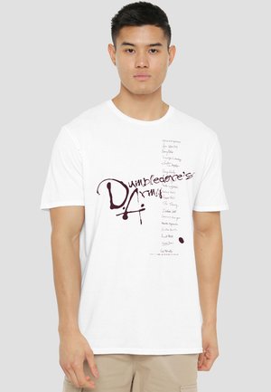 Young man wearing a white T-shirt with "Dumbledore's Army" text and graphic design, standing against a plain light gray background.