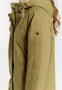 Olivengrøn parka-jakke med hætte, der har lynlås, snoretræk og logo-badge på ærmet. Tekstureret, vandafvisende stof.