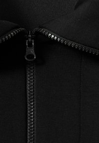 Indumento zip nero con una texture liscia. Presenta una zip centrale con denti in plastica e un tirante, circondata da un colletto ripiegato.
