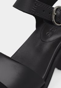 Μαύρο δερμάτινο σανδάλι με φαρδιά μπροστινή λωρίδα, λουράκι στον αστράγαλο με ασημένια αγκράφα και χαμηλό τακούνι τύπου block, σε κοντινή λήψη.