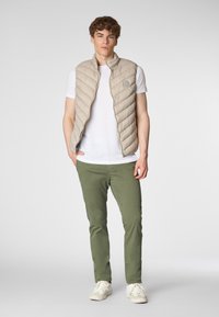 Un giovane uomo sta in piedi con sicurezza, indossando un gilet imbottito beige sopra una maglietta bianca e pantaloni verde oliva, con sneakers bianche.
