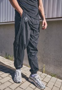 Black, lightweight joggers with a relaxed fit and elastic ankles, featuring a circular logo on the left hip. Worn with gray athletic shoes.