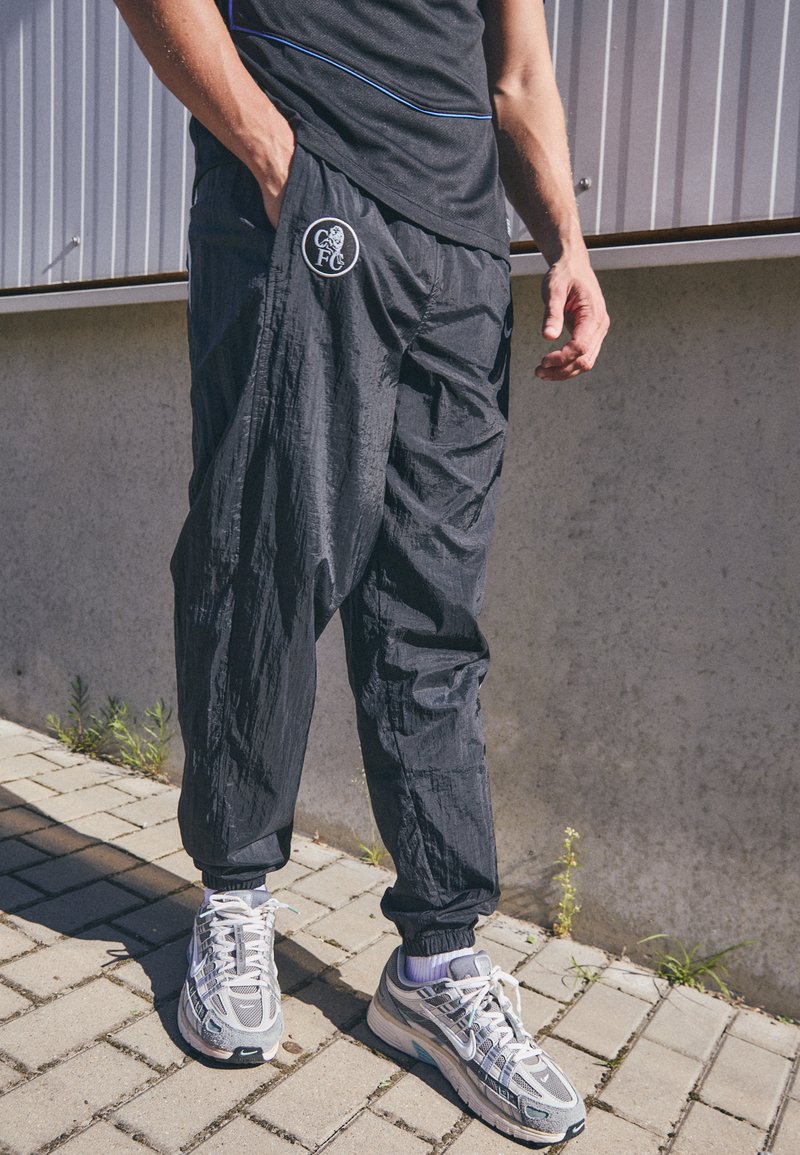 Jogging léger noir avec une coupe décontractée et des chevilles élastiques, arborant un logo circulaire sur la hanche gauche. Portés avec des chaussures de sport grises.