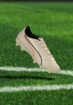 Chaussure de football blanche avec des accents noirs et rouges flottant au-dessus de l'herbe verte et des lignes blanches du terrain.