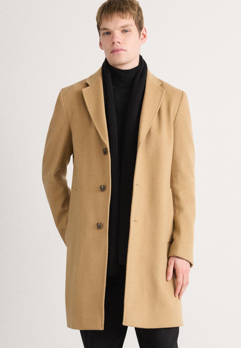 Beige wool overcoat with a notched collar, four brown buttons, and side pockets, worn over a black turtleneck and dark trousers.