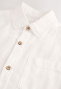 Weißes Leinenhemd mit klassischem Kragen, drei Holzknöpfen und einer aufgesetzten Tasche auf der linken Seite. Strukturierter, leichter Stoff.