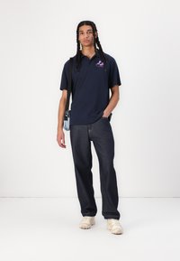Navy polo shirt with logo, paired with dark blue jeans and cream sneakers. Model carries a small light blue crossbody bag.