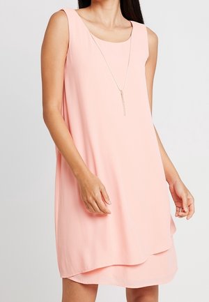 Femme portant une robe sans manches rose clair à volants et un long collier en argent, debout devant un fond uni.