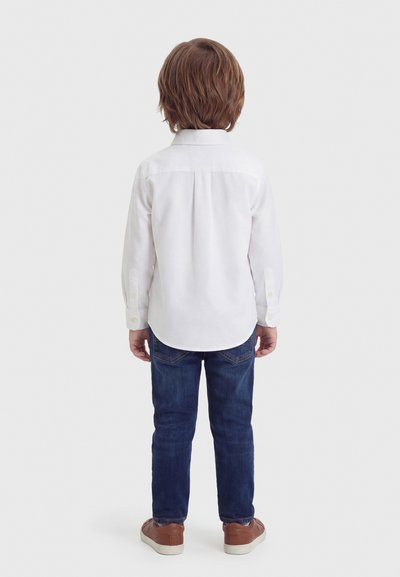 Παιδί με μεσαίο καστανό μαλλί που φοράει λευκό μπλουζάκι μακρυμάνικο, σκουρόμπλε τζιν και καφέ αθλητικά παπούτσια, στέκεται γυρισμένο με την πλάτη σε λευκό φόντο.