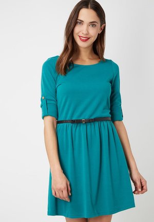 Mujer sonriendo, vestida con un vestido de teal hasta la rodilla, con mangas de tres cuartos enrolladas y un delgado cinturón negro en la cintura, contra un fondo liso.