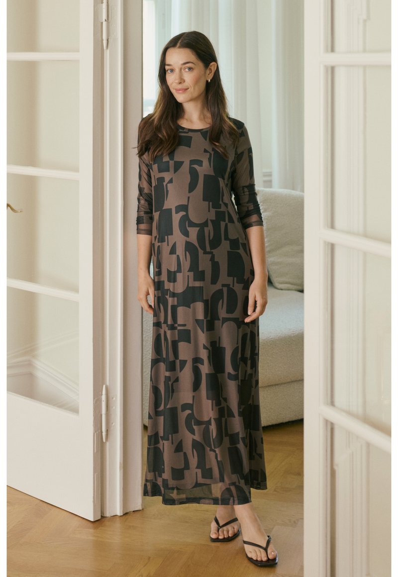 Maxikjole i et gennemsigtigt brunt stof med abstrakte sorte geometriske mønstre, med lange ærmer og en afslappet pasform, kombineret med sorte sandaler.