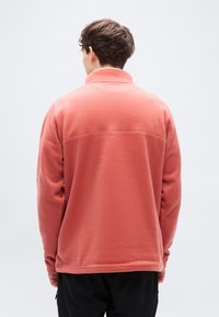 Πουλόβερ από κοραλλί φλις με ψηλό γιακά, υφασμάτινη υφή και ίσια λάστιχα, με ραφή στο πάνω μέρος της πλάτης.