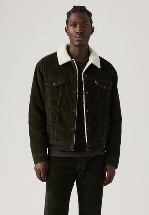 Homme portant une veste en velours côtelé vert foncé avec un col en peau lainée blanche et un pantalon assorti, debout devant un fond clair uni.