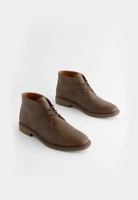 Botas de tobillo de cuero marrón con una textura suave, punta redonda y diseño tradicional de cordones. Presentan una suela cosida en contraste y un tacón robusto.