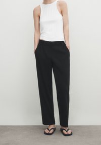 Pantaloni neri a gamba larga con vita alta, tasche laterali e una texture di tessuto liscia, abbinati a una canotta bianca a costine. Infradito nere.