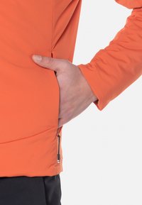 Jasnopomarańczowa kurtka wykonana z gładkiego materiału. Posiada boczną kieszeń z zamkiem błyskawicznym oraz dopasowany krój rękawów. Zbliżenie na dłoń w kieszeni.