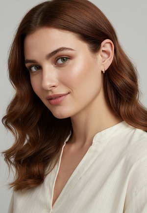 Orecchini a cerchio dorati adornano una donna con capelli mossi castano chiaro. Indossa una camicia color crema con colletto e sottili righe verticali.