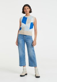 Knit sleeveless sweater featuring a geometric pattern in shades of blue and beige, with a ribbed neckline and hem, paired with wide-leg jeans.