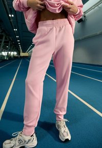 Heledat roosad dressipüksid, mis on valmistatud pehmest kangast, varustatud elastse vöökohaga ja kitsenevate mansettidega, millel on valged spordijalatsid sinisel rajal.