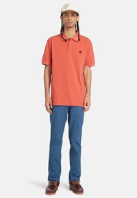 Orange polo skjorta med kort krage, marinblå byxor och bruna skor. Klädseln har en liten logotyp på skjortan och strukturerad tyg.