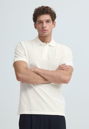 Młody mężczyzna z kręconymi włosami, ubrany w białą koszulkę polo i ciemne spodnie, stoi z założonymi rękami na tle jasnej, jednolitej ściany.