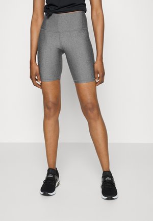 Person iført grå tætsiddende knælange shorts, sort bundet crop top og sorte atletikskor, stående mod en ensfarvet baggrund.