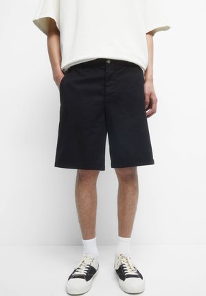 Sorte skræddersyede shorts lavet af bomuld med lige snit, der har sidelommer og knaplukning. Bæres med hvide strømper og sneakers.