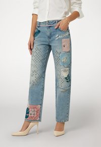 Šviesiai mėlynos džinsinės kelnės su rožinėmis ir mėlynomis aplikacijomis, turinčiomis gėlių ir geometrinių raštų, pažeistą tekstūrą ir tiesų kirpimą.