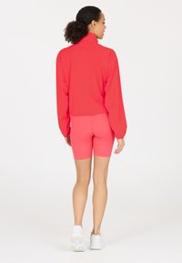 Bright pink athletic jacket with a high collar, long sleeves, and a cropped design. Paired with matching shorts. White sneakers.
