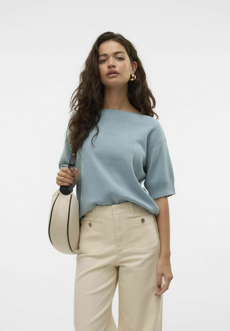 Suéter de punto azul claro con escote barco y mangas cortas, combinado con pantalones de cintura alta en color crema. La modelo sostiene un bolso beige con detalles en negro.