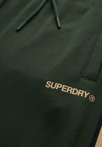 Mørkegrøn sweatshirt med snoretræk, prydet med et guld "SUPERDRY" logo. Glat tekstur og enkelt, funktionelt design med sidelommer.
