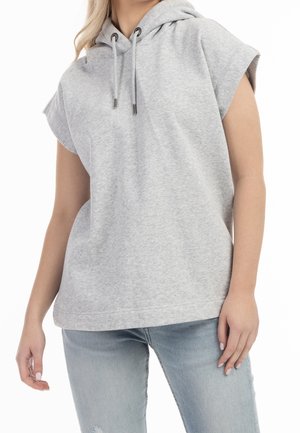 Femme portant un sweat à capuche sans manches gris clair et un jean bleu clair avec une déchirure au niveau d'un genou sur fond blanc.