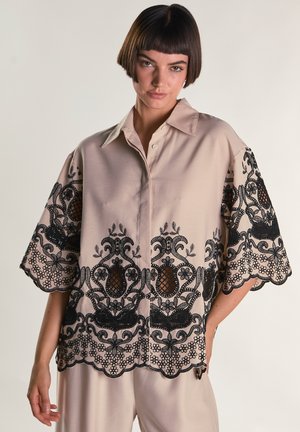 Frau mit kurzen dunklen Haaren, die eine beige Bluse mit weiten Ärmeln trägt, welche am Saum und an den Manschetten eine kunstvolle schwarze Spitzenstickerei aufweist.