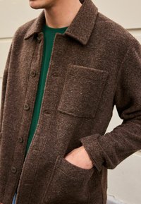 Chemise en laine marron avec un col, fermeture à boutons, poche avant et poches latérales, présentant une finition texturée et une coupe décontractée.