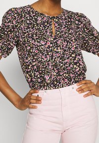 Blouse noire à fleurs avec manches ballon, détail d'ouverture en forme de goutte devant et taille froncée. Associée à un jean rose clair taille haute.